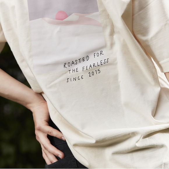 Obrys oblaků s růžovým sluncem nad nimi a nápis „ROASTED FOR THE FEARLESS SINCE 2015“ na zadní straně bílého trička.